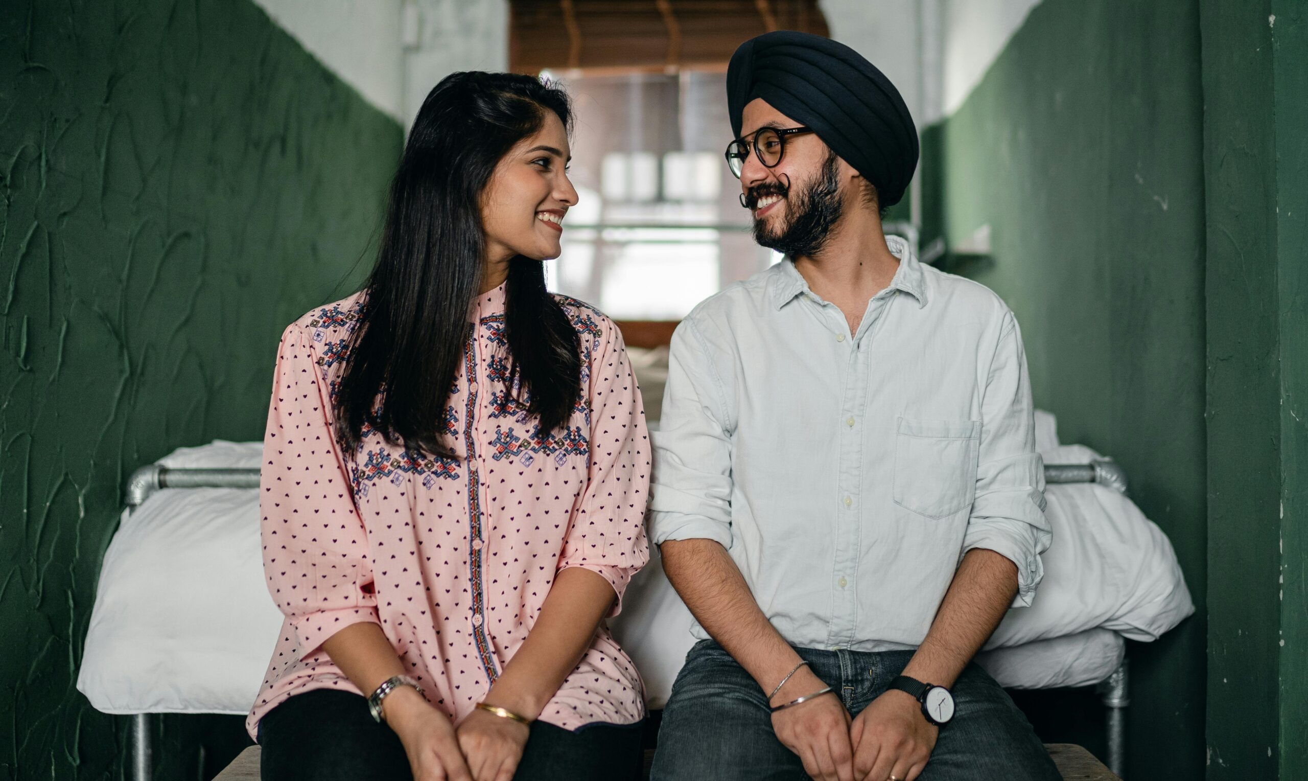 A happy couple sitting on a bed, looking at each other affectionately in a green-walled room.